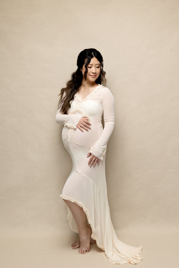 Pregnant woman in a long, flowing cream dress poses against a beige backdrop, gently holding her belly.