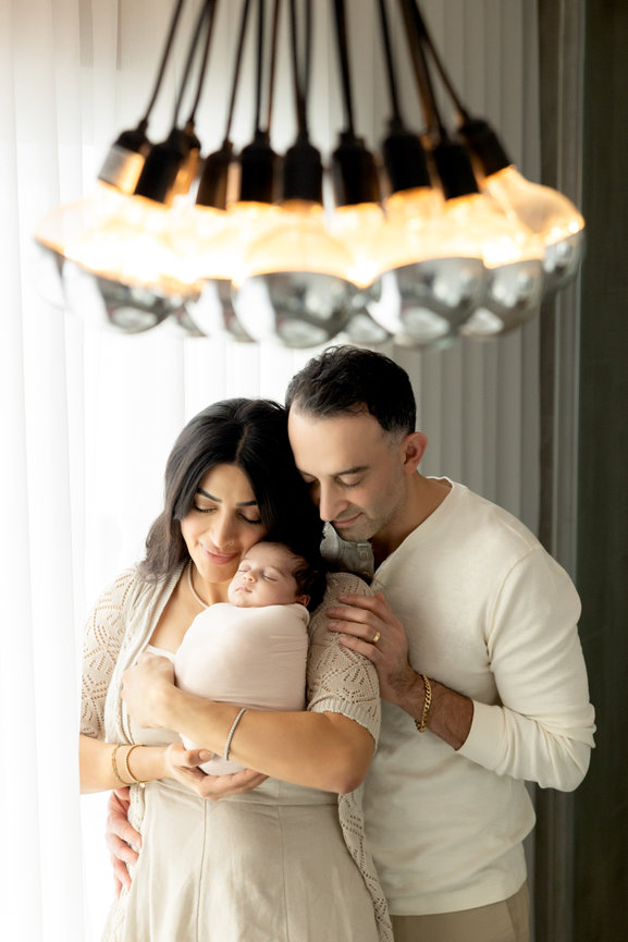 A couple lovingly embraces their swaddled newborn under a warm, modern chandelier.