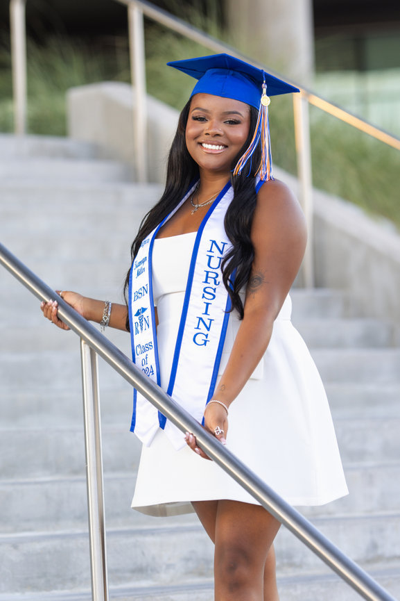 UTA Graduate in a white dress and blue cap gown smiling on stairs.
