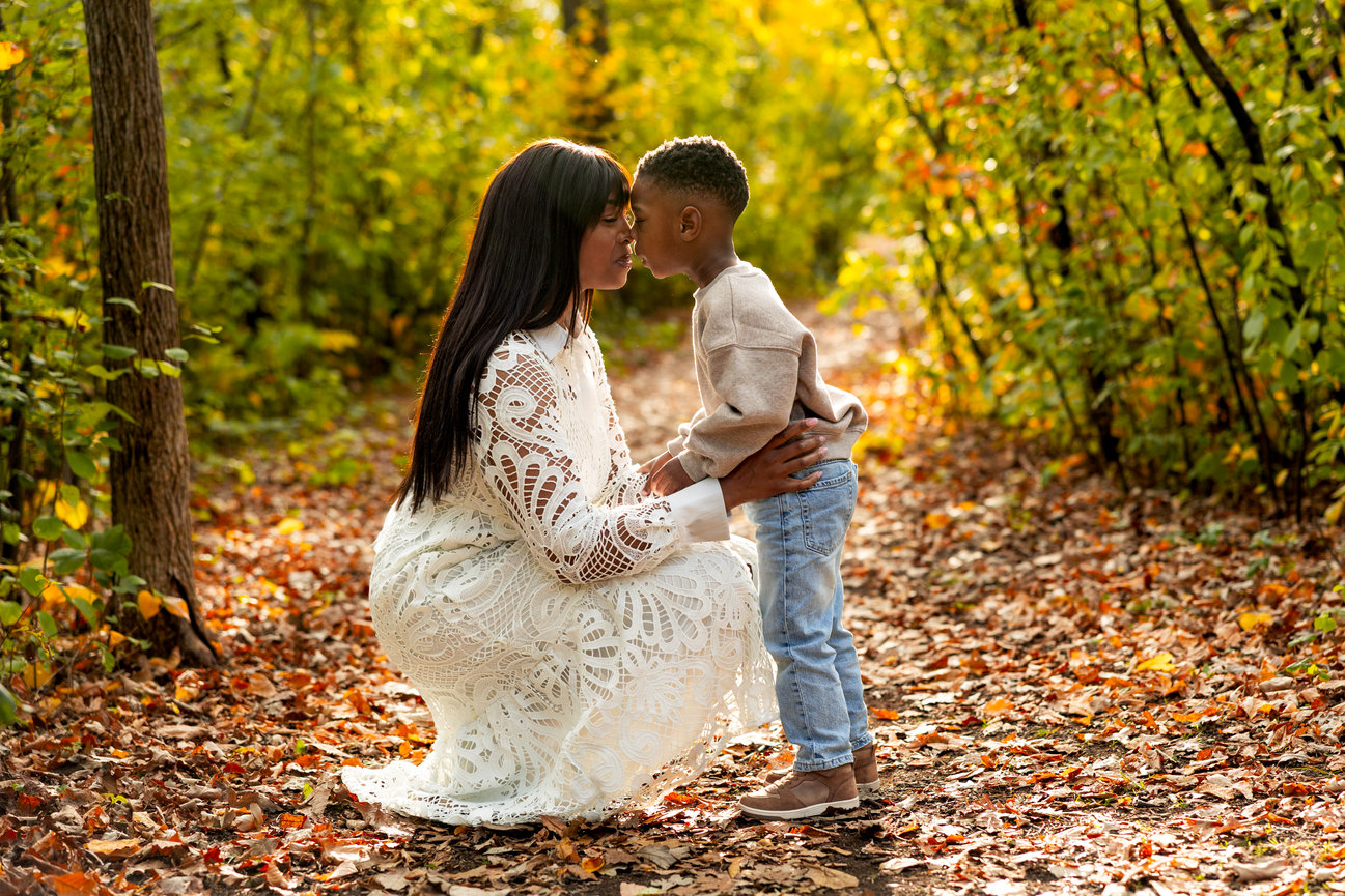 Woman in white dress kneeling on leaf-covered path, gently touching noses with a child in a forest setting.
