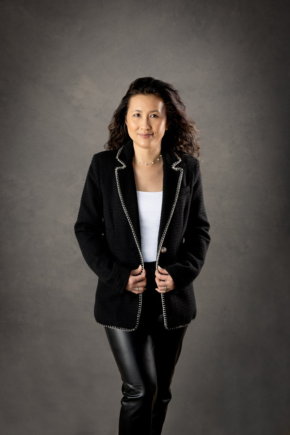 Woman in a black blazer and leather pants standing against a gray backdrop, smiling confidently.