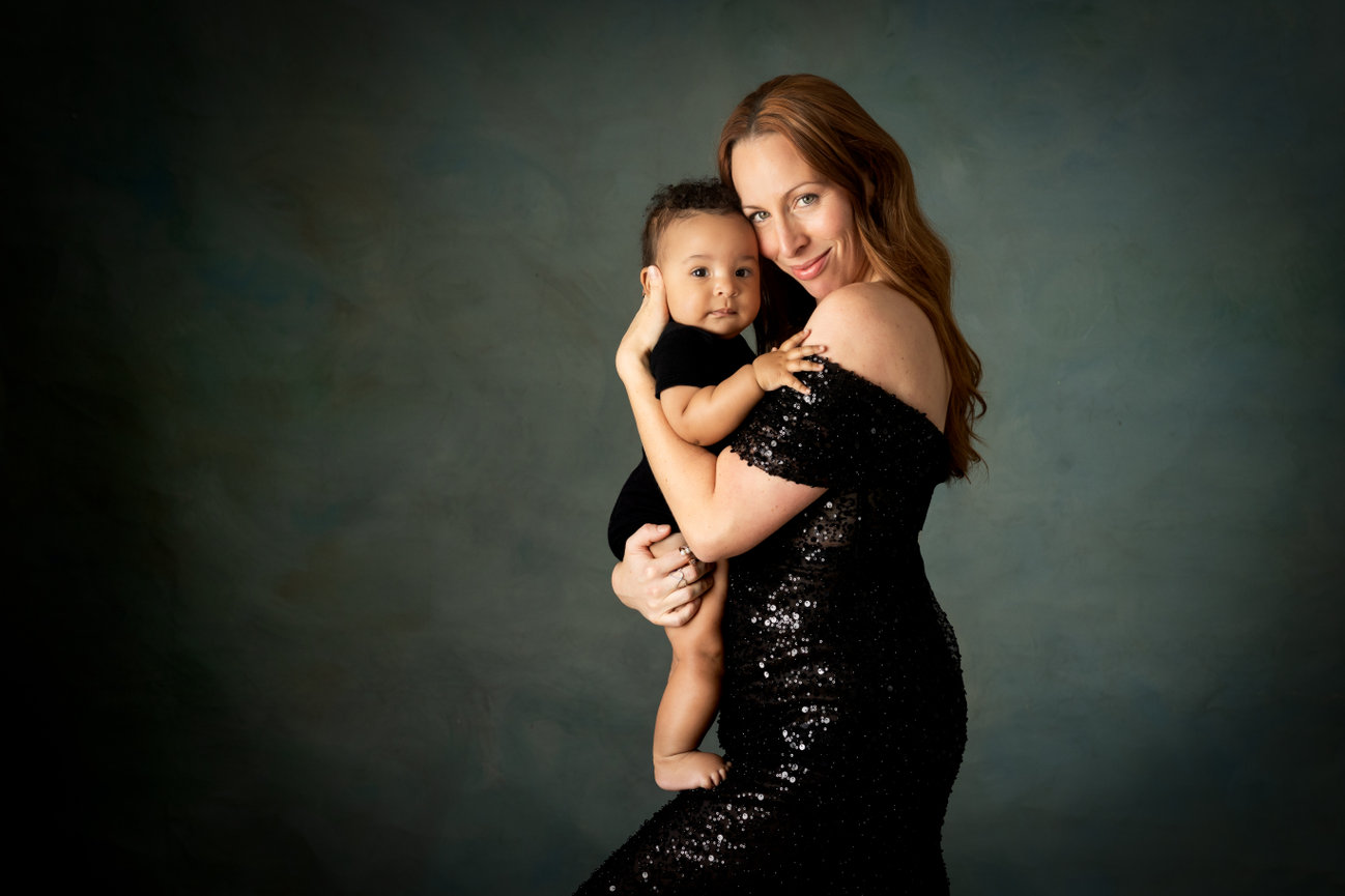 Woman in a black dress holds a baby against a gray-blue background.