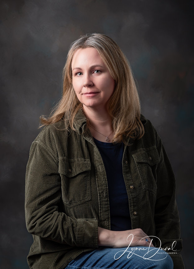 Blonde woman wearing a green jacket, jeans, and black shirt sitting down