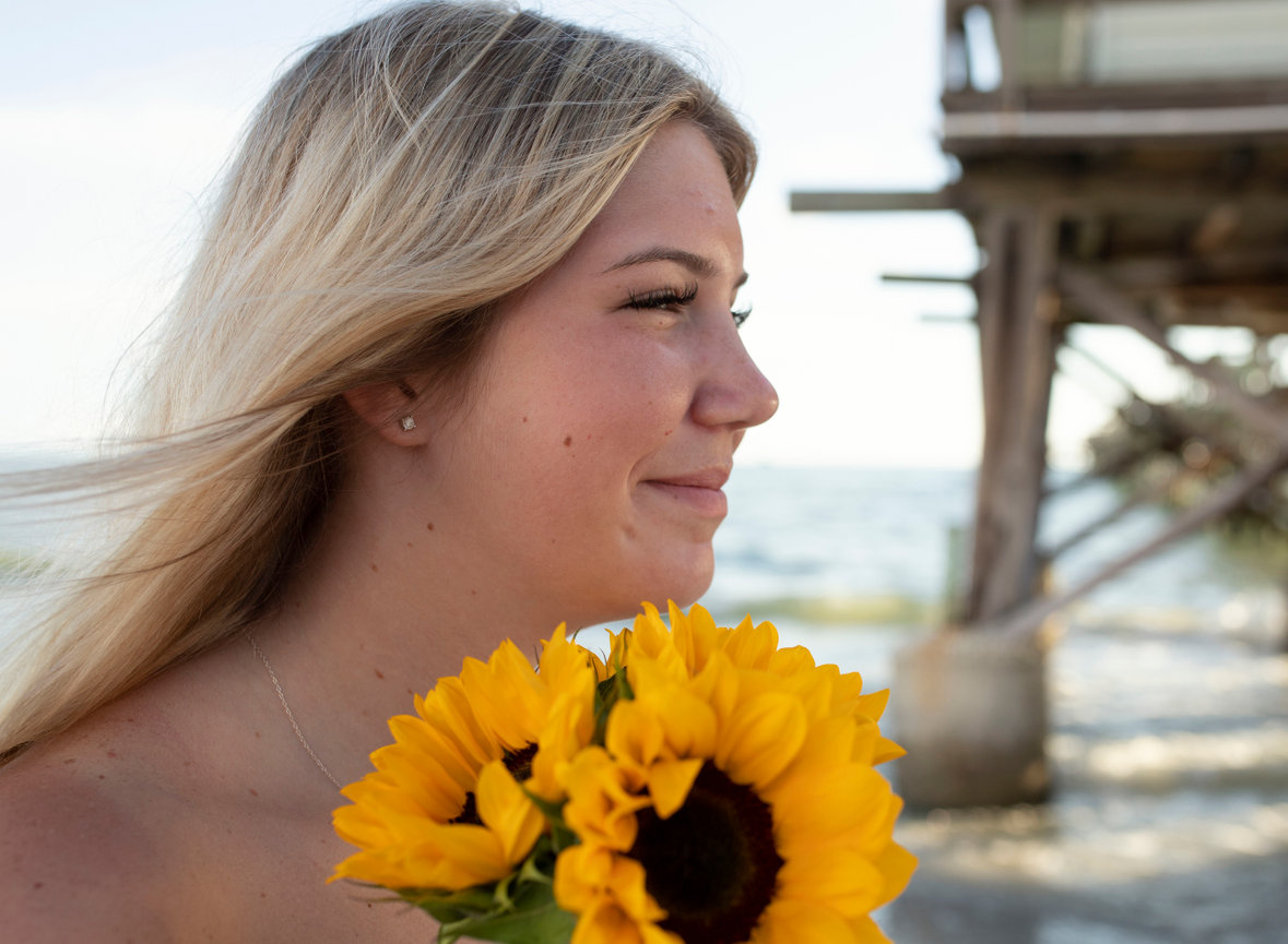 Senior girl holding sunflowers at the beach during for her destination portrait session with Angie Vincent of Heart to Art Photography.