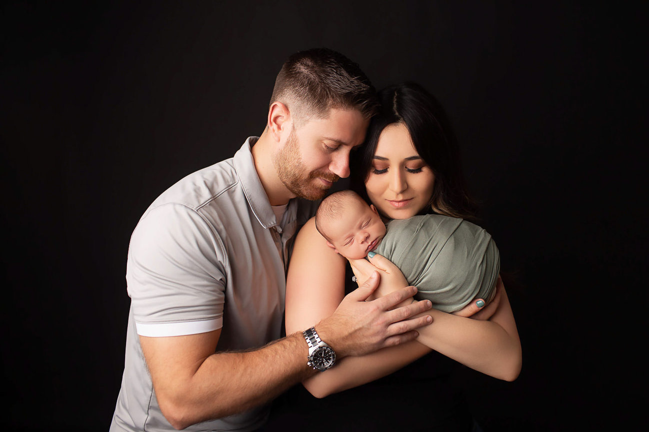 Newborn baby wrapped in sage fabric sleeping in moms arms with dad intimately hugging mom during newborn photoshoot in Kansas City with Dreamworks Photography 