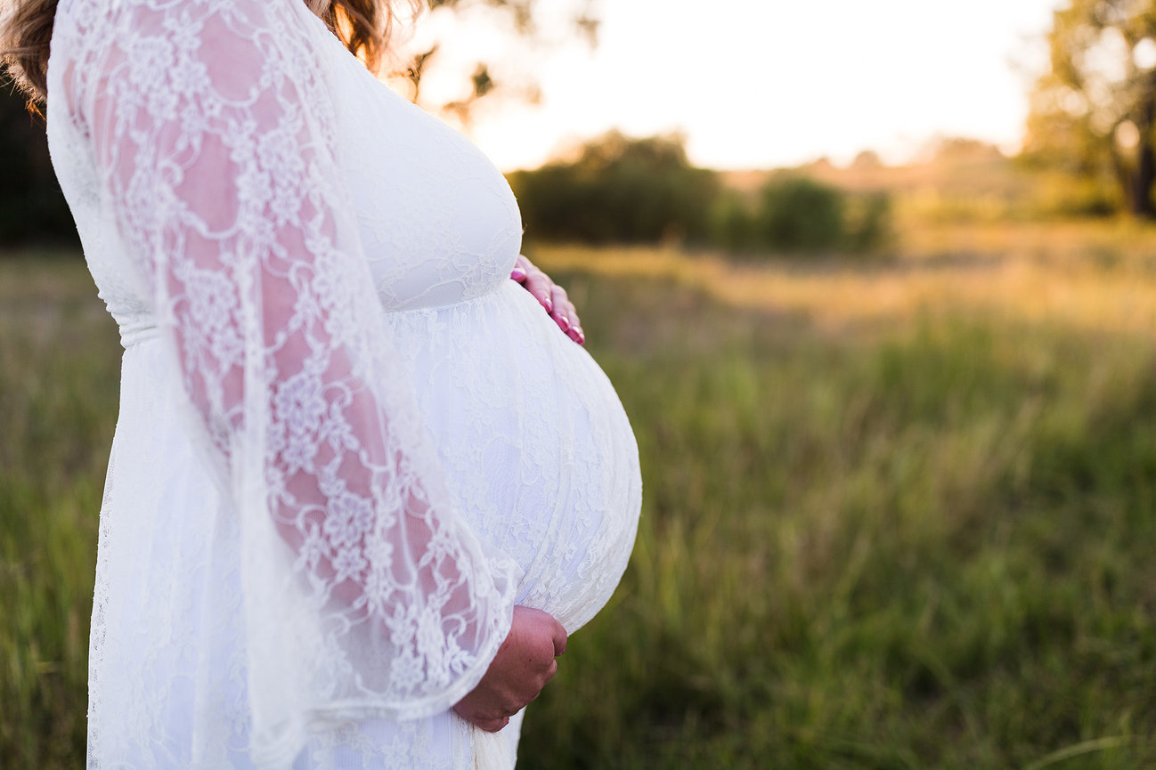Pregnant woman in white lace dress standing in a grassy field at sunset.
