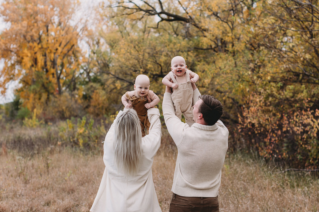 Bennington Family Photographer captures couple lifting two smiling babies in a field with autumn trees.