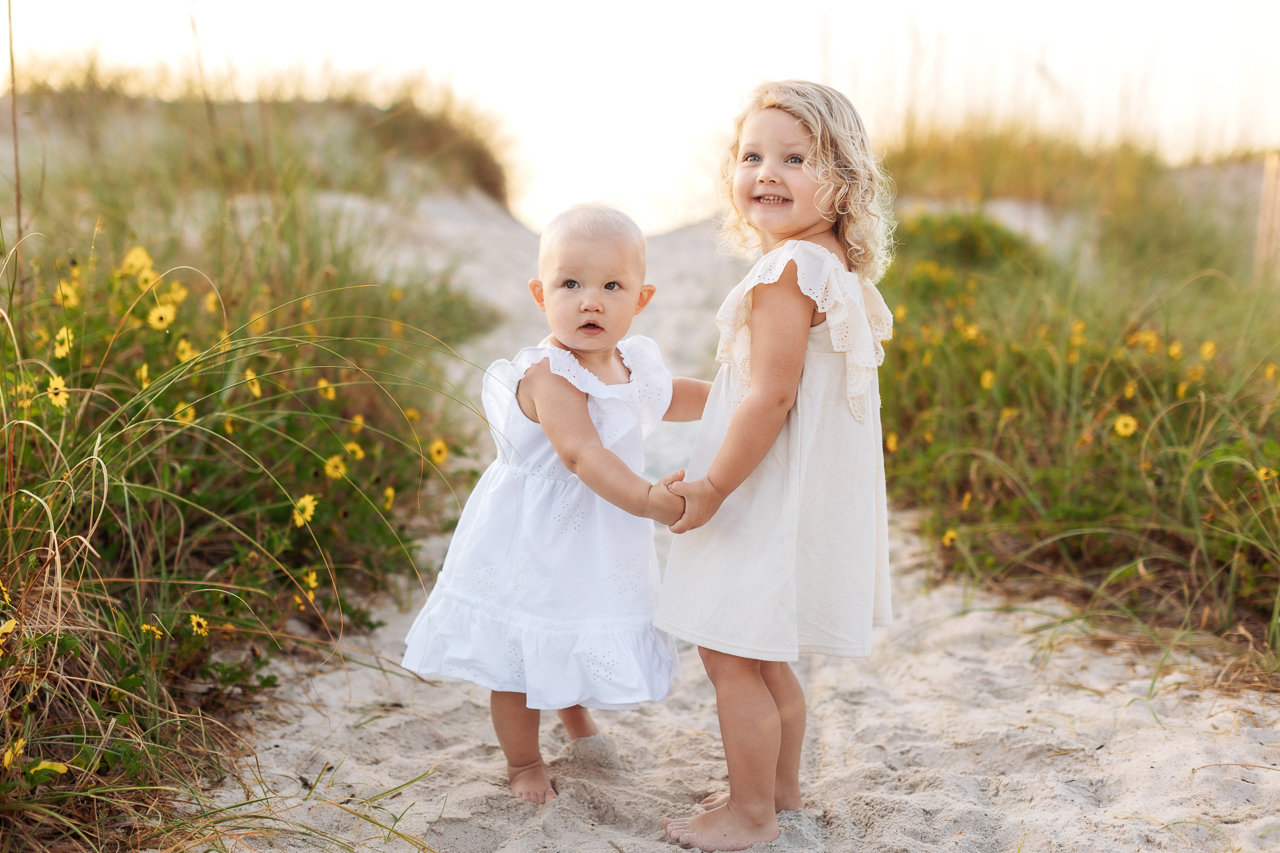 Two young children in white dresses stand on a sandy path surrounded by grass and wildflowers.