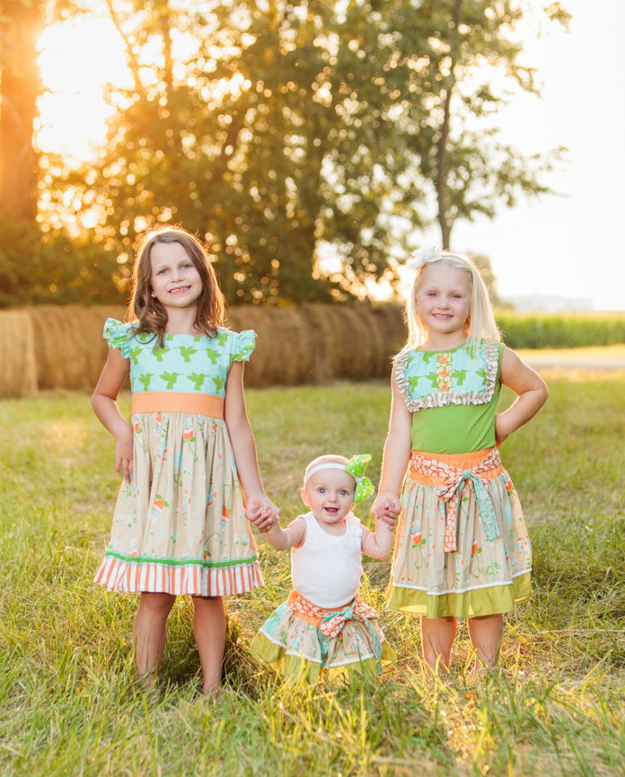 Three children holding hands and smiling outdoors, wearing matching dresses, with hay bales and trees in the sunny background.