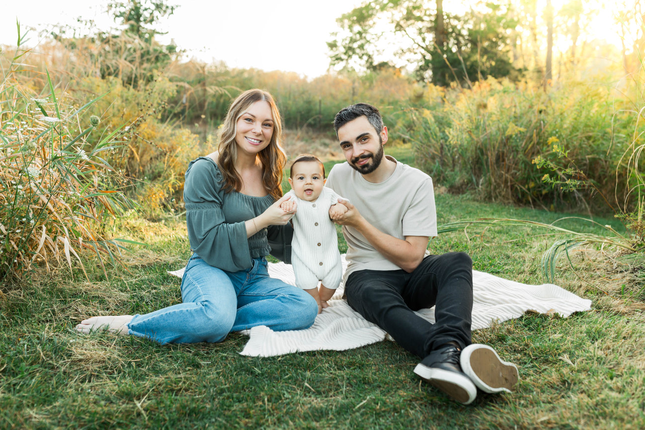 Family of three with baby seated on blanket during outdoor session in Farmington, MI