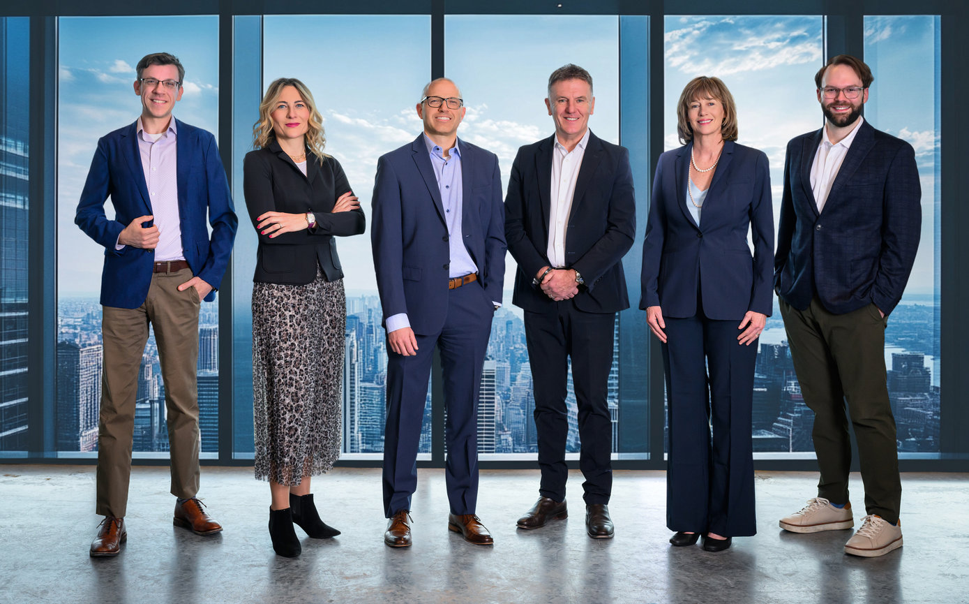A group of six professionals in business attire, posing in front of large windows with a city skyline backdrop.