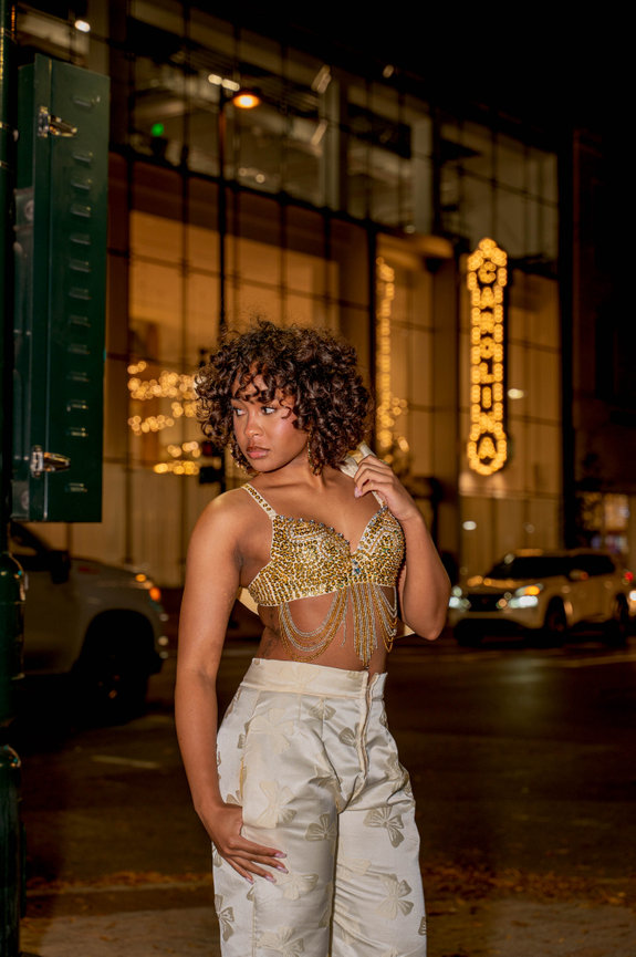 A person with curly hair poses confidently in stylish attire on a nighttime city street with illuminated signage behind captured in the style of Jo Hayes Images Charlotte Portrait Fashion Photographer Charlotte Event Photography Discovery Place