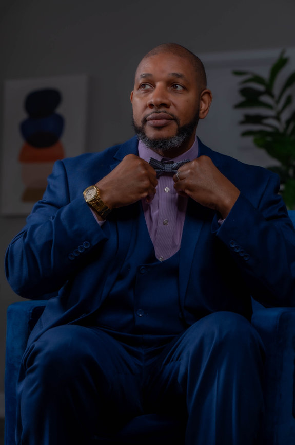 Man in a blue suit adjusting his bow tie, seated, with an abstract painting and plant in the background in an editorial style captured by jo hayes images charlotte portrait photographer
