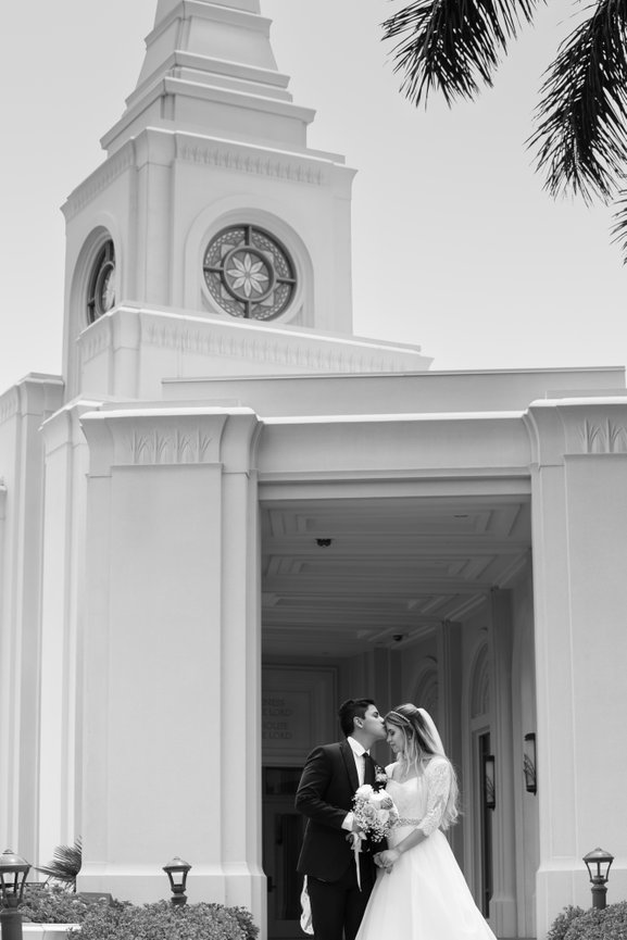 Bride and groom kissing in front of a church building with a prominent tower, surrounded by greenery.