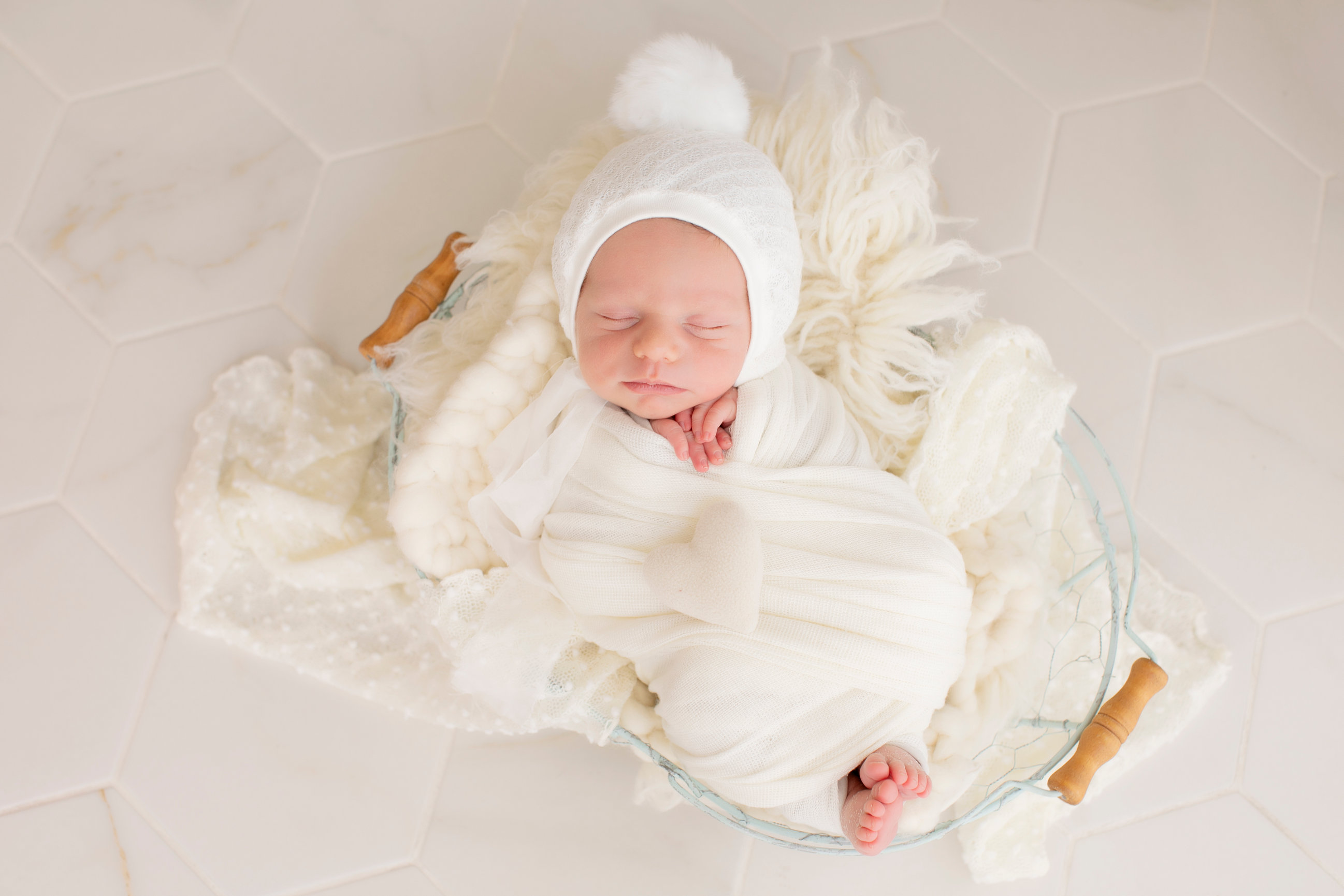 Sleeping newborn wrapped in ivory, resting in a wooden bowl on a cream blanket.