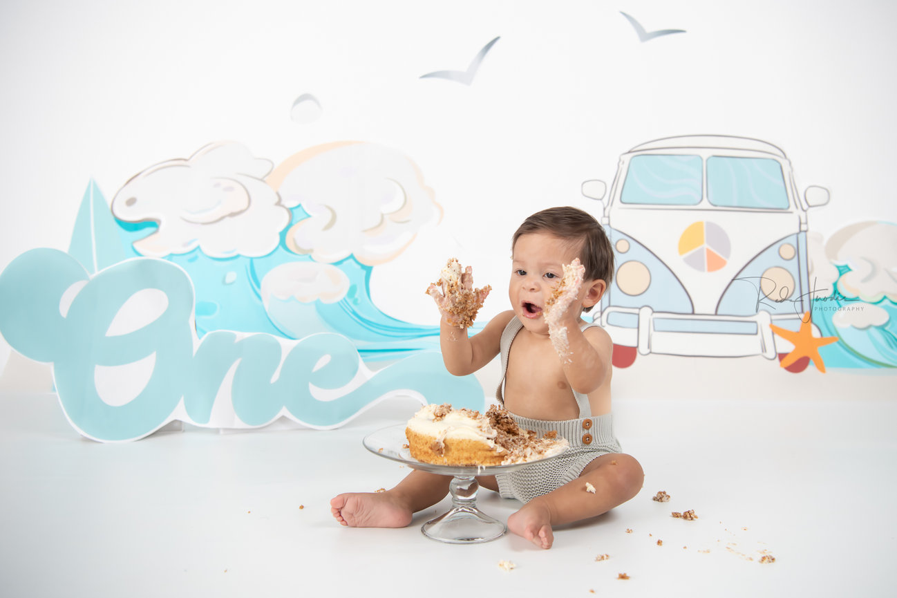 A joyful baby sitting on the floor, enjoying a cake smash with frosting on hands, surrounded by a beach-themed backdrop.