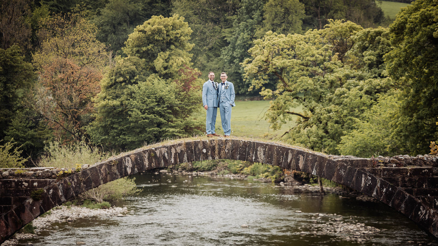 Two grooms in light blue suits standing on a stone bridge over a river in a lush forest.