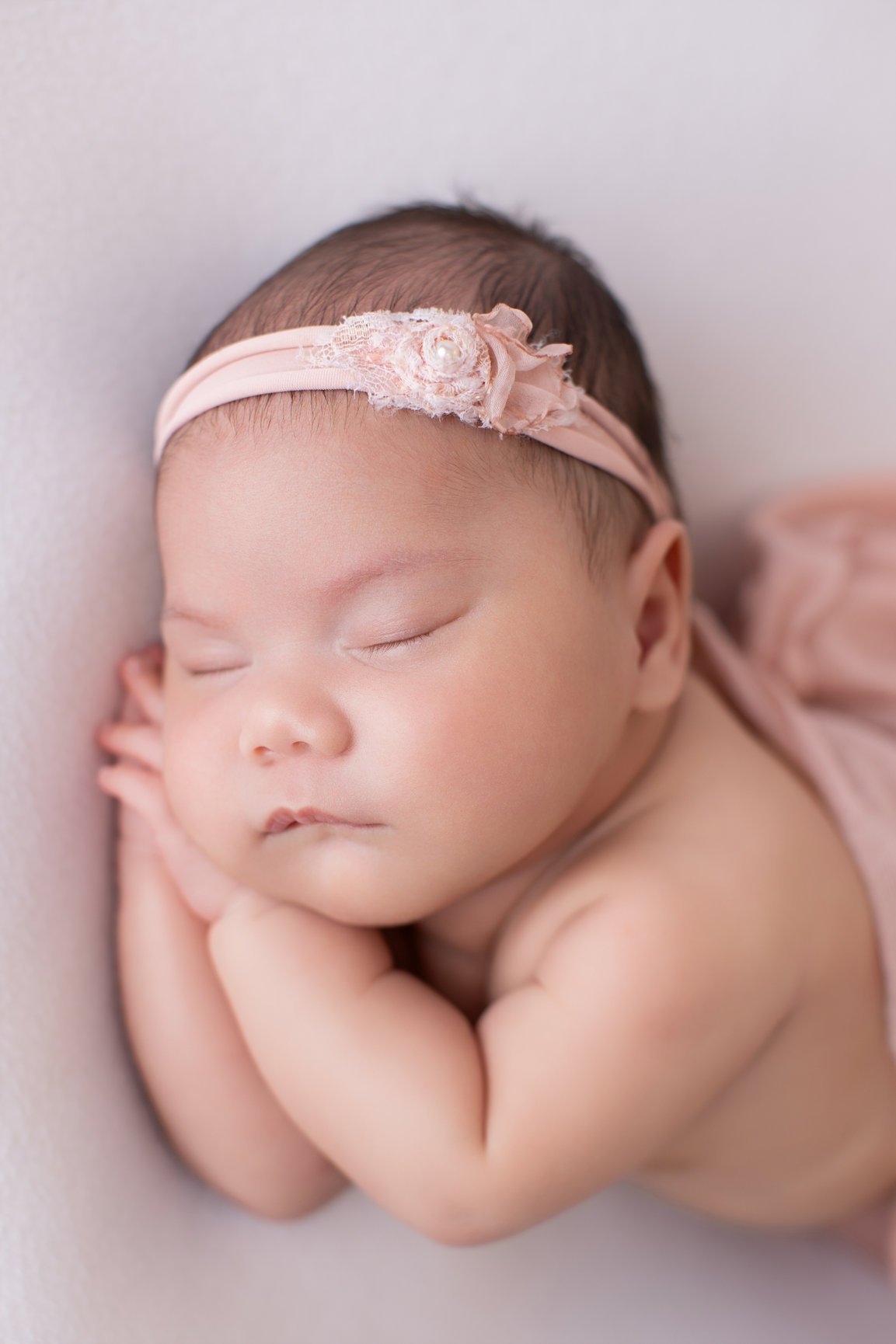 Close-up of a sleeping newborn girl resting on her hands in a blush pink headband
