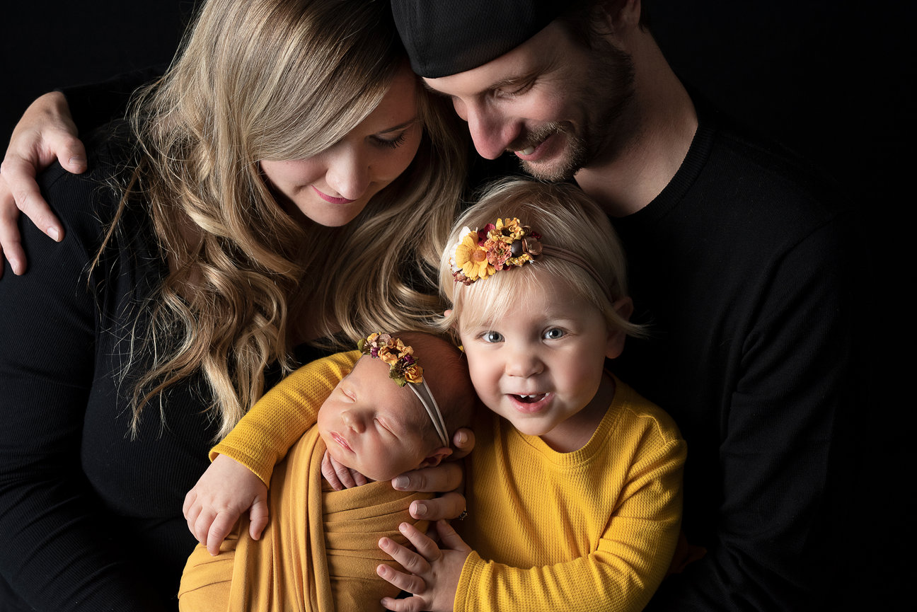 A loving family portrait in Fox River Grove, Illinois features a mother, father, a toddler, and a newborn snuggled in a mustard-colored blanket. The children wear matching floral headbands. The smiling family is dressed in coordinating outfits.