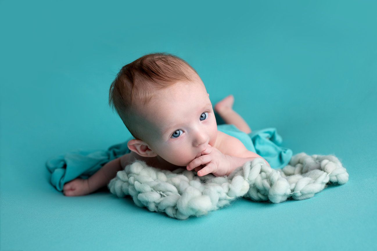 A three-month-old baby with light hair and blue eyes lies on a fluffy, knitted cream blanket against a turquoise background. Clad in turquoise clothing, the little one has one hand near their mouth, gazing upward with curiosity.