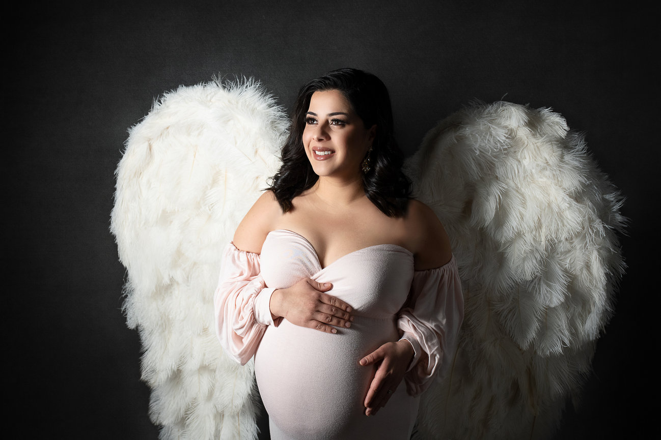 A pregnant woman stands against a dark background, wearing a light pink, off-the-shoulder dress. She is smiling and has large white feather wings on her back. Her hands rest gently on her belly.