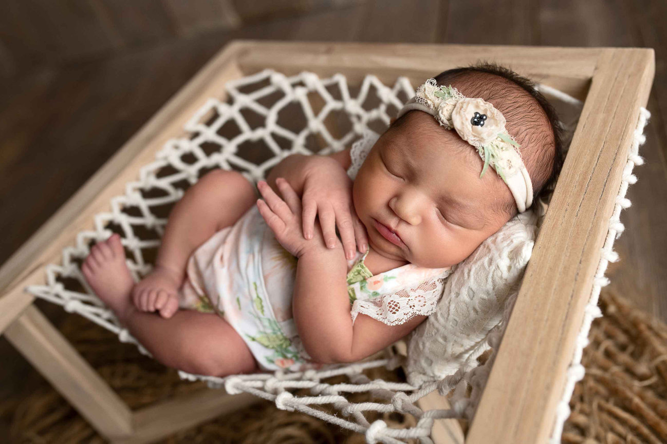 A newborn baby sleeps peacefully in a small hammock, with hands gently clasped. Dressed in a floral outfit and matching headband, the baby rests within this serene scene in Plato Center, Illinois. The hammock, supported by a wooden frame.