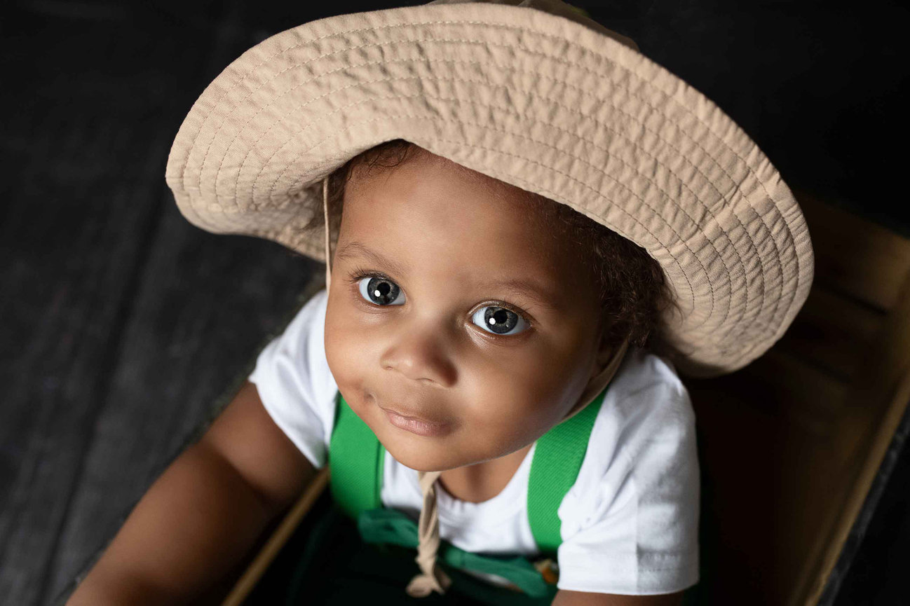 A child with large eyes is wearing a wide-brimmed hat and a white shirt with green suspenders. Their gentle expression gazes upwards, reminiscent of the serene landscapes of Schaumburg, Illinois, set against a dark background.