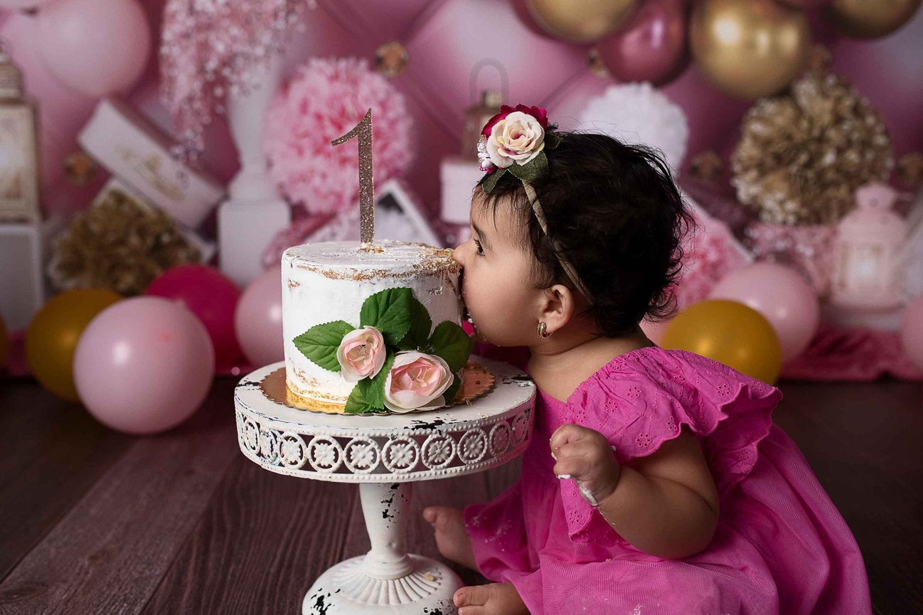 In Arlington Heights, a baby in a pink dress leans forward to take a bite of a small, decorative cake with a 1 topper. The cake is adorned with roses, while colorful balloons and flowers set the scene for this joyful Illinois birthday celebration.
