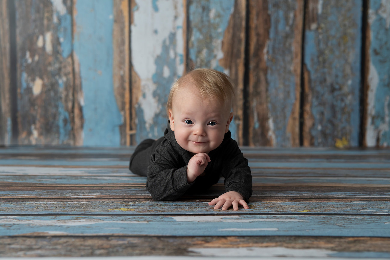 A baby photo captures a light-haired infant in a dark outfit, lying on their stomach on a wooden floor. With a blue and brown rustic background, the baby looks toward the camera, smiling brightly.