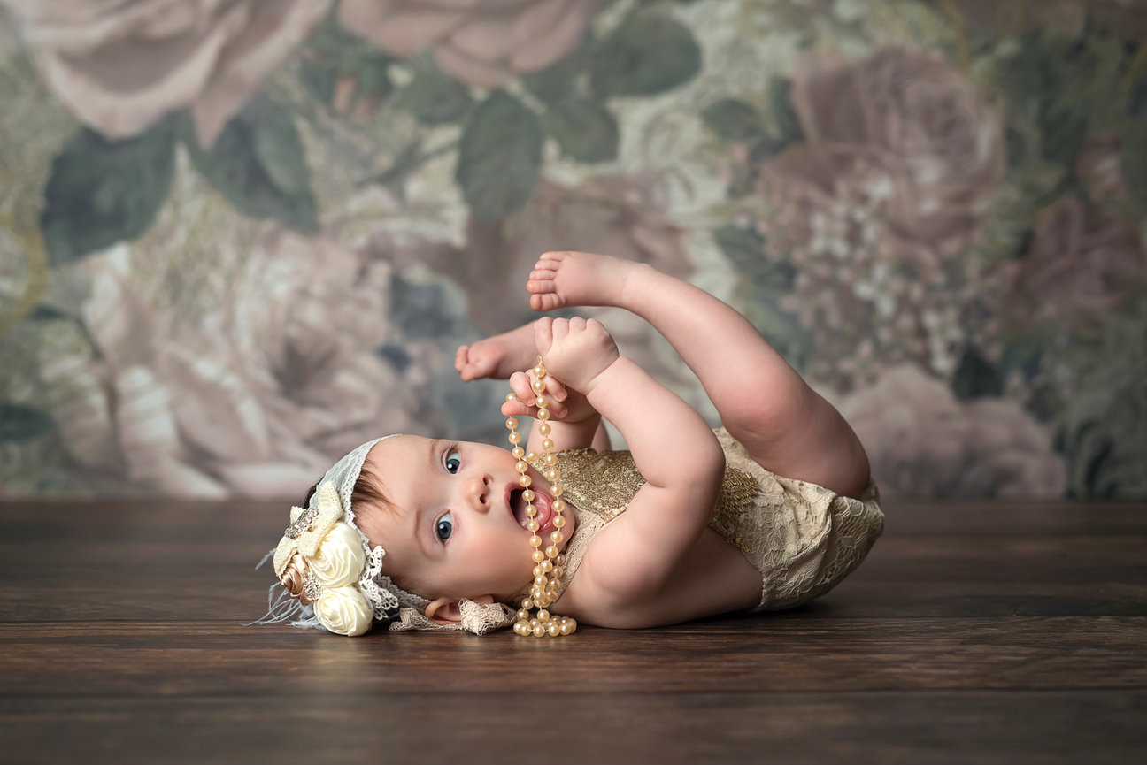 A baby in a cream-colored outfit and floral headband lies on a wooden floor, playing with a pearl necklace. Captured in a photo studio near me, the background features large, soft floral patterns in muted tones.
