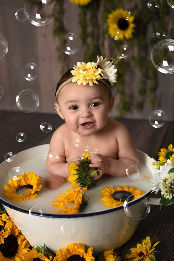 The baby wears a sunflower headband, holds a sunflower, and giggles as bubbles float around. The background, adorned with hanging greenery and sunflowers, creates a charming scene perfect for child photography near me.