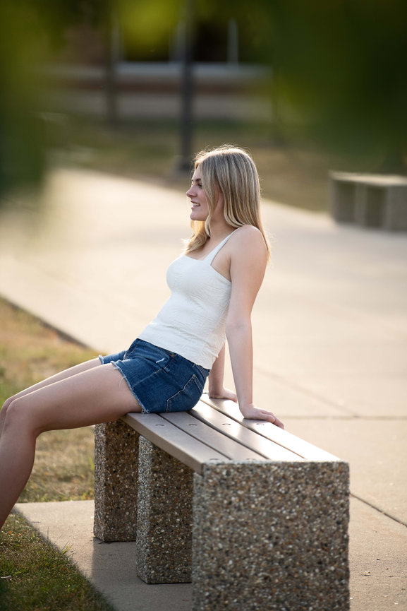 A woman with long blonde hair sits relaxed on a park bench, capturing the essence of senior photography. She wears a white tank top and denim shorts, her hands resting behind her. The background is a paved path and grassy area, softly lit by sunlight
