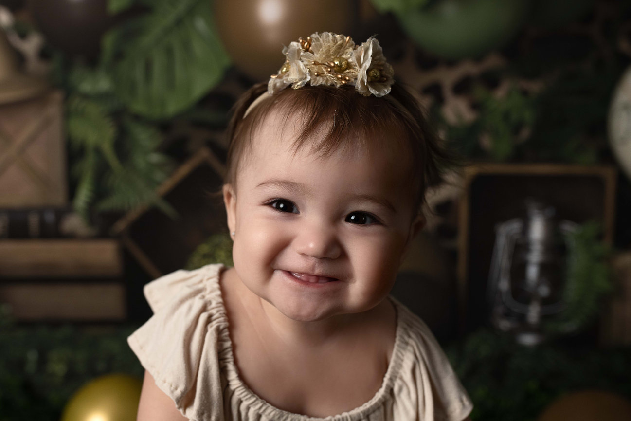 A smiling toddler wearing a small flower headband and a light-colored dress is set against a leafy backdrop with earthy brown and green tones.