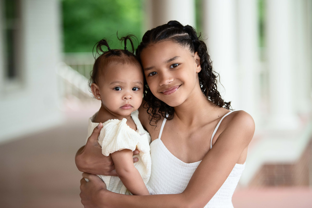 A young girl with curly hair smiles warmly as she holds her curious baby sibling. Both are dressed in light-colored clothing, standing on a porch adorned with white pillars and lush greenery in the background.
