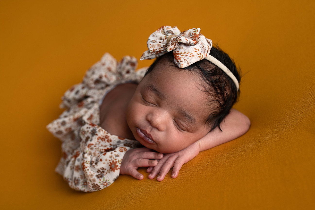 A sleeping African American newborn baby with a floral headband rests on a mustard-colored surface. The baby wears a matching floral outfit and is posed with hands gently under their cheek, creating a peaceful scene.