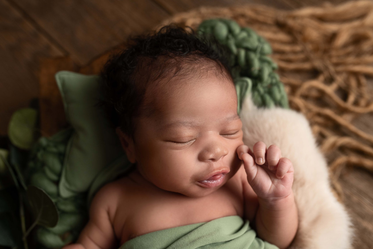 A sleeping baby wrapped in a green blanket lies peacefully on a soft surface in Bull Valley. The baby, with curly hair and a serene expression, appears content. The background features a textured, natural woven material that enhances the scene.