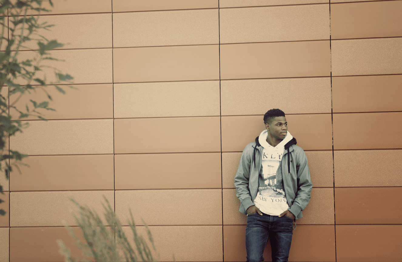 An African American teenage guy wearing a graphic hoodie stands against an orange tile wall.