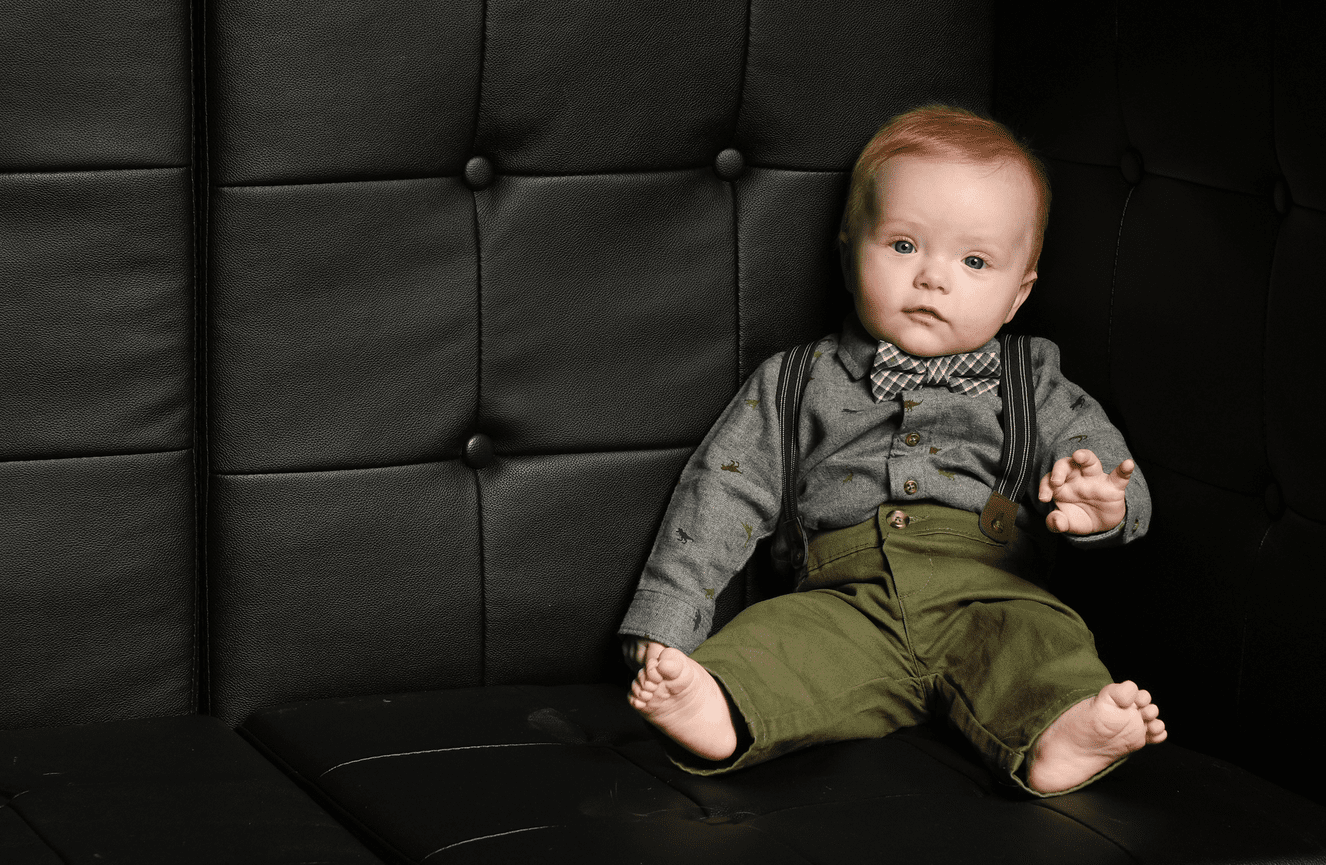 A redheaded toddler boy wearing green trousers and suspenders shows off his bare toes at Kliks Photography.