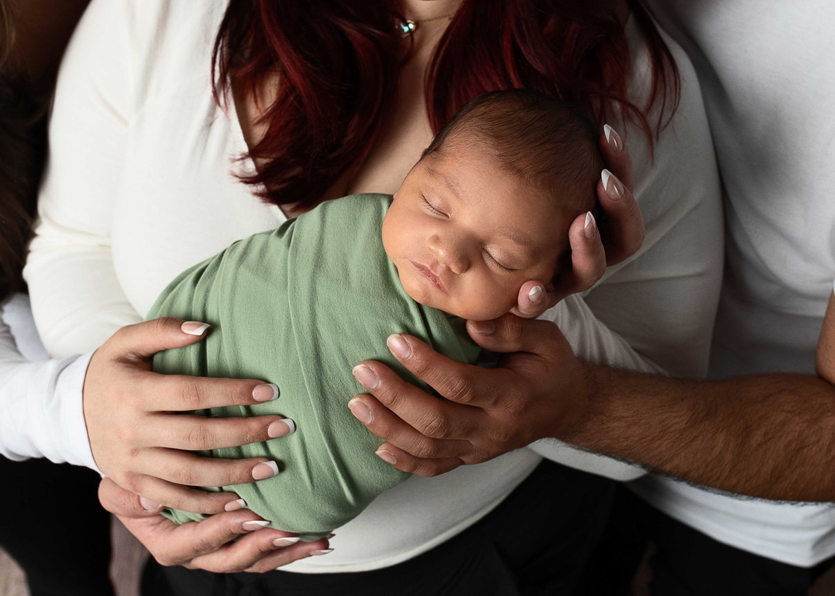 Newborn baby boy wrapped in green swaddle, cradled lovingly in parents' hands – newborn photography in Streamwood, Illinois