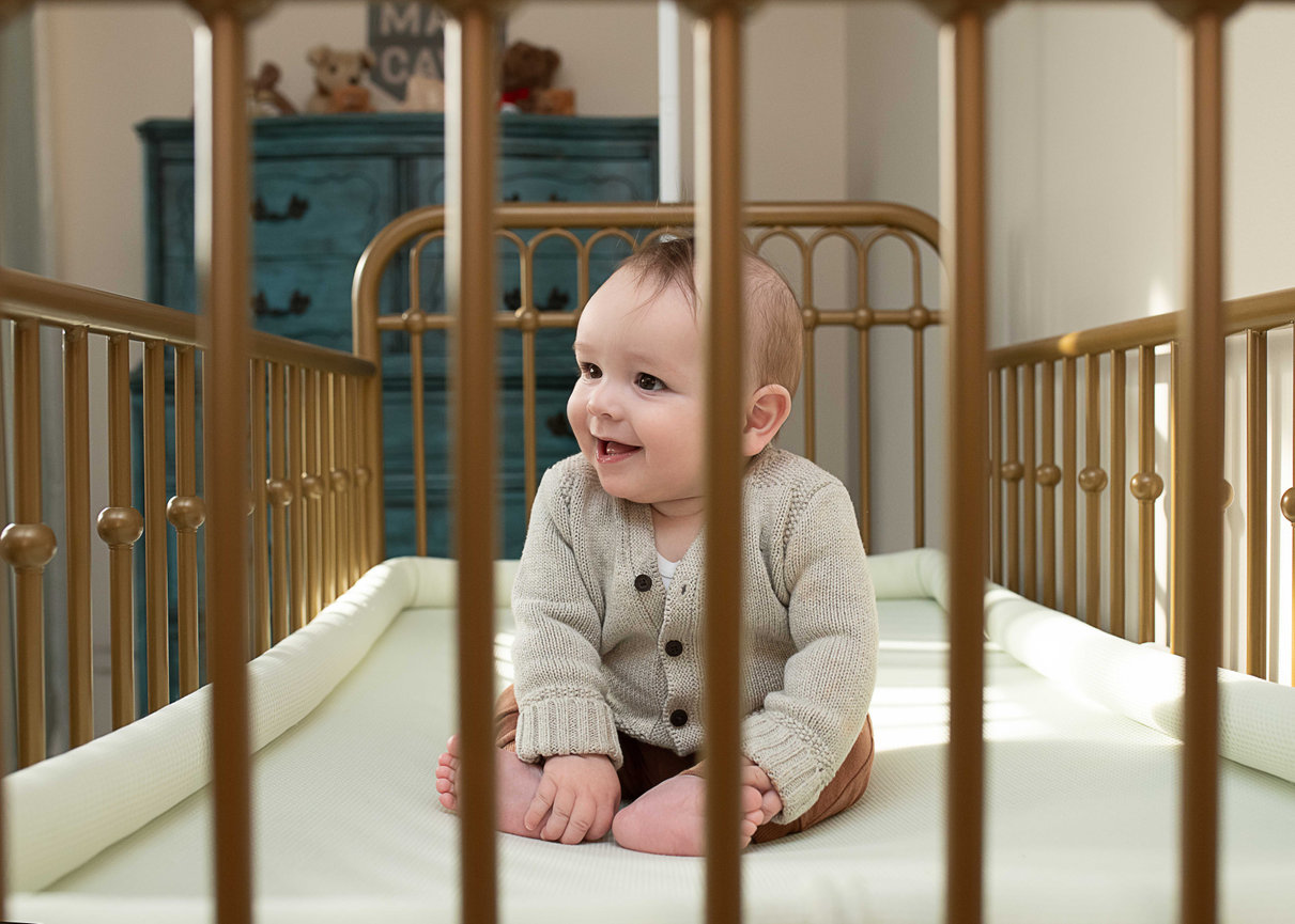 Smiling baby sitting in a brass crib with vertical bars, wearing a knit sweater.