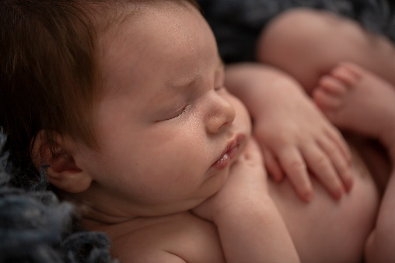 Clean, simple close up naturally posed newborn portrait of a baby boy with chubby cheeks. One hand is tucked beneath his chin while the other rests on his belly. Image is dramatically short lit with soft lighting to create a cozy and intimate feel.