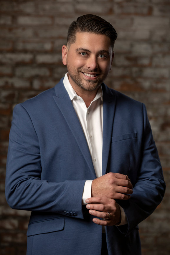Striking modern business portrait headshot of a professional in a dress shirt and jacket with a brick wall as a backdrop.