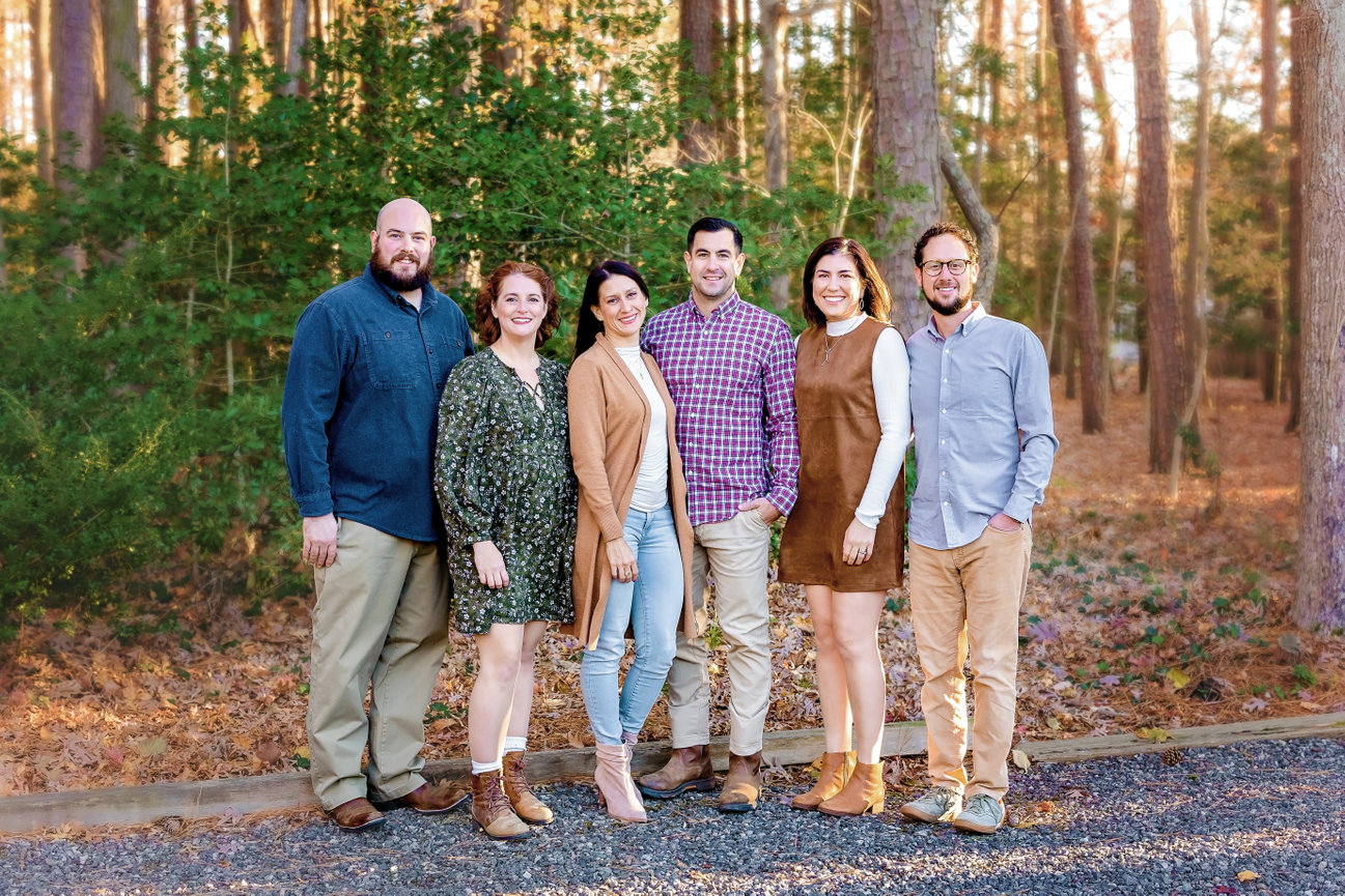 Outdoor photography session of a group of six adults smiling outdoors in a wooded area with casual attire.
