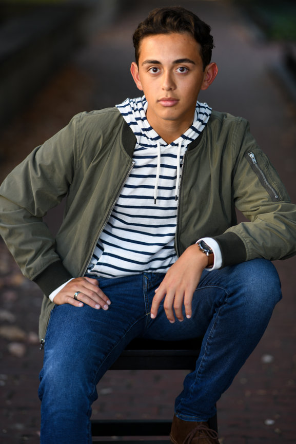 Teenage boy with an army green jacket, striped shirt and jeans sitting for senior photos in Dublin OH