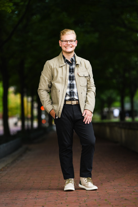 Teenage boy in a tan jacket, plaid shirt and jeans standing under a dark canopy of trees for senior pictures in Columbus OH.