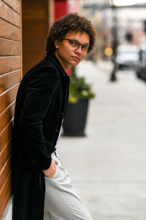 Teenage boy in a long black velvet jacket and white pants leaning against a brick wall for senior portraits in Dublin OH.
