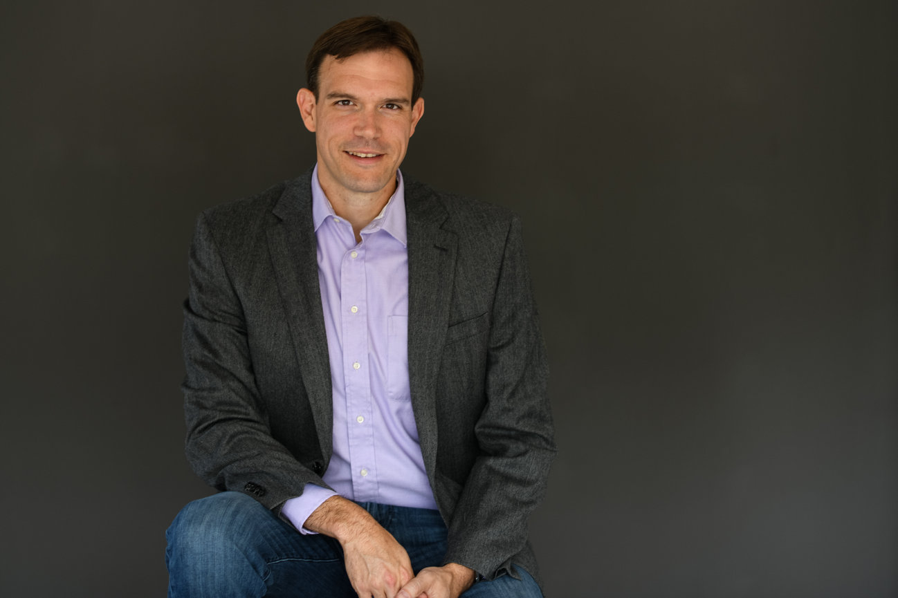 Man in a gray blazer and purple shirt seated against a dark background, smiling confidently for professional branding photos in dublin ohio boutique studio.