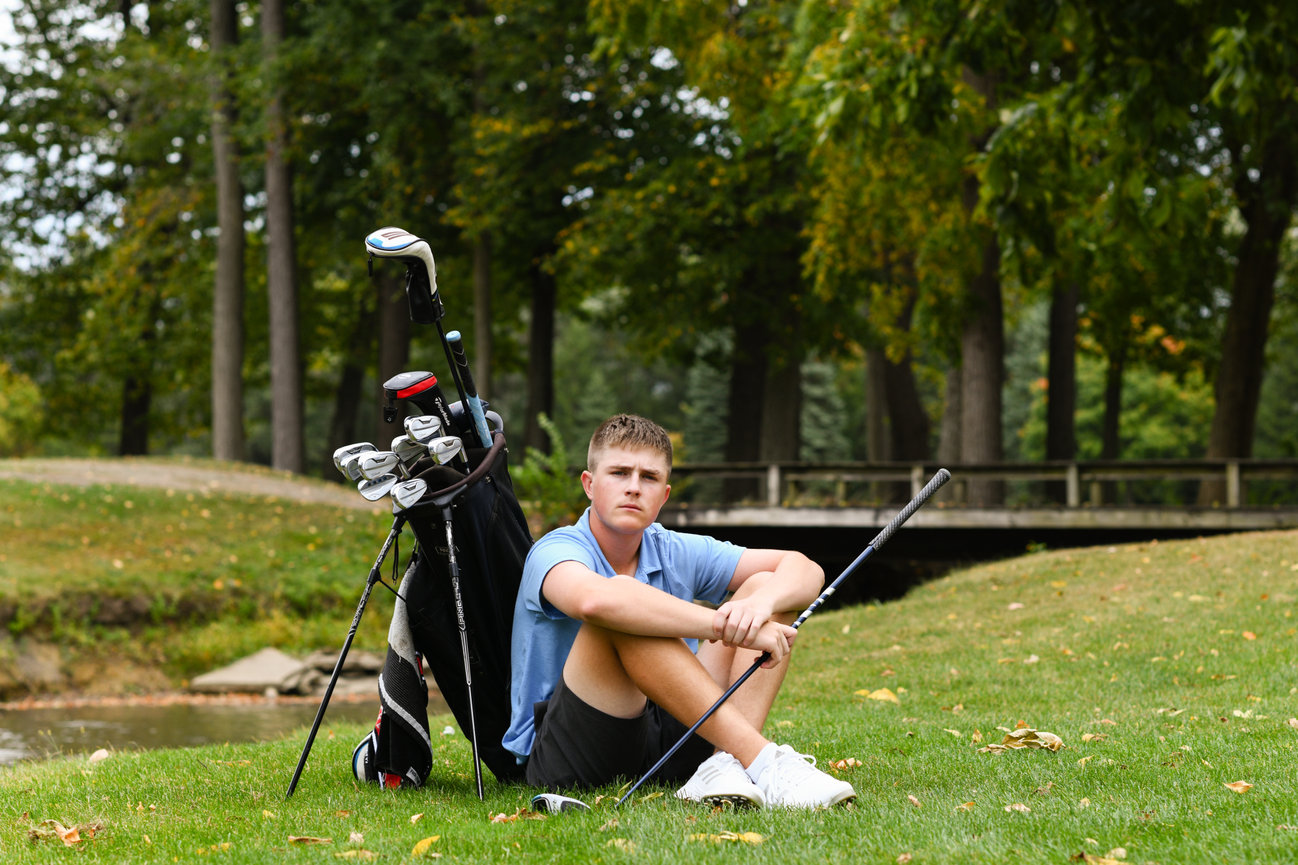 Teen golfer sitting on the grass, leaning against a golf bag with clubs, in a park setting.