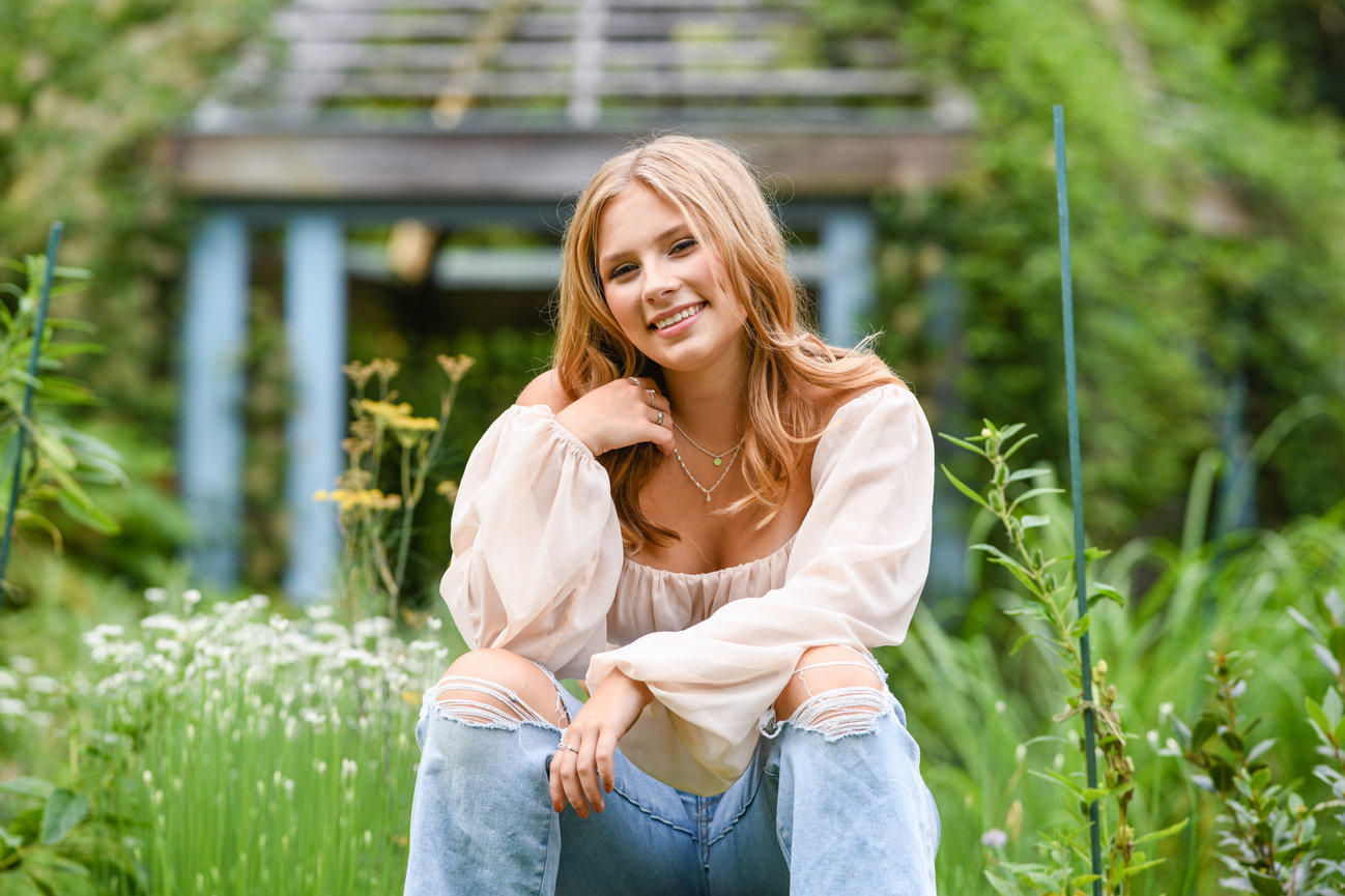 Teen girl sitting on a stone wall wearing distressed denim and a soft flowy blouse in a lush garden setting