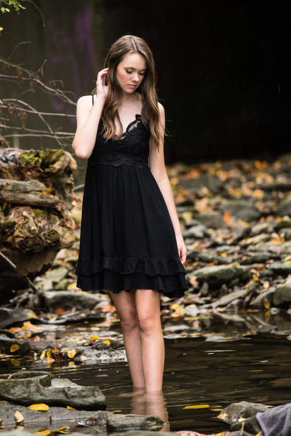 Teenage girl in a black lace dress walking through a pool of water in the forest for senior pictures in Dublin OH.